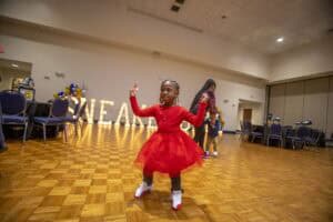 A young girl in a red dress dances joyfully on a wooden floor, surrounded by festive decorations.