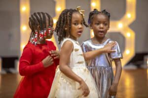 Three young girls in festive dresses engage playfully together, with colorful hair accessories.
