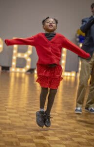 Jumping girl in red outfit at CAES News event with illuminated letters in the background.