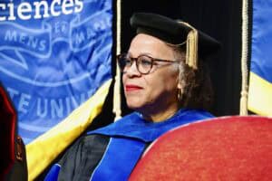 A woman in academic regalia smiles during a graduation ceremony, with banners in the background.