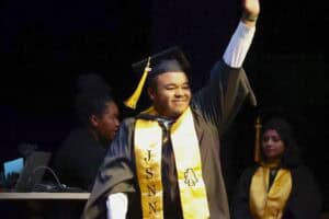 A graduating student in cap and gown waves to the audience, smiling proudly. Other graduates stand nearby.