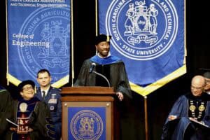 A speaker in academic regalia addressing a graduation ceremony, with university banners in the background.