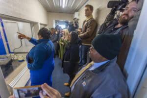 A woman in blue points while a crowd of journalists and onlookers observes through a window.