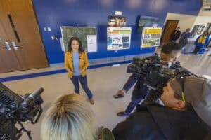 A woman speaks to cameras in a blue-walled hall, with posters and an audience in the background.
