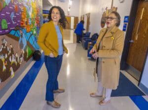 Two women converse in a brightly lit hallway with a colorful mural behind them. One wears a mustard jacket.