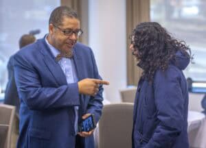 A man in a blue suit and bow tie talks and points playfully to a woman wearing a navy jacket in a meeting room.