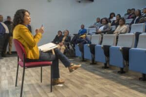 A woman in a yellow jacket speaks to an engaged audience in a lecture setting.