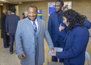 A smiling man in a gray coat interacts playfully with a woman while another man looks on in a casual setting.