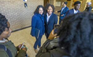 Two women smile for a photo indoors, surrounded by others in business attire. A person takes their picture from behind.