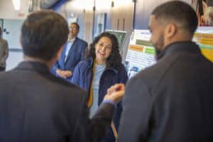 A woman in a blue jacket smiles while engaging with two men during a discussion in a hallway.