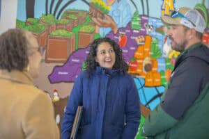 Three people engage in conversation in front of a colorful mural depicting agriculture and various food items.