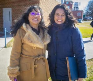Two women smile while standing outdoors. One wears a brown coat with a fur collar, the other a dark blue jacket.