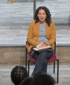 A woman with curly hair sits on a chair, smiling while holding papers, engaged in a discussion with others.