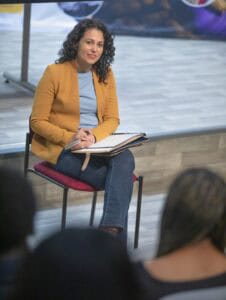 A woman with curly hair sits on a chair, holding a notebook and looking thoughtfully at an audience.