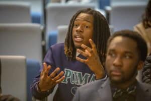 A young man passionately discusses a topic while gesturing, seated among others in an auditorium setting.