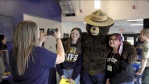 CAES News logo with people taking photos of a bear mascot wearing a cowboy hat.