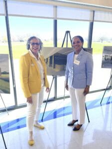 Two women stand beside presentation boards in a bright space, smiling. One wears a yellow blazer, the other a light…