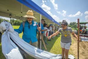 A group of people set up a tent at an outdoor event, with one person holding a tarp and another pointing.