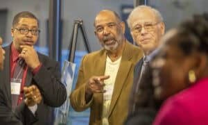 Three men engaged in conversation, expressing various emotions and gestures, with blurred figures in the background.