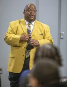 A speaker in a bright yellow suit addresses an audience, holding a microphone and gesturing.