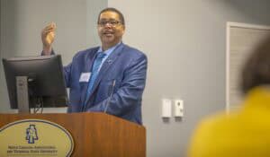 A speaker gestures while presenting at a podium with a university logo. Audience members are visible in the background.