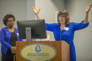 A woman in a blue dress gestures while speaking at a podium, with another woman listening attentively nearby.