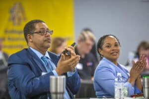 Two attendees engaged at a conference, one clapping, the other smiling, with a yellow banner in the background.