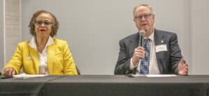 Two speakers sit at a table during a discussion, with one in a yellow jacket and the other in a suit.