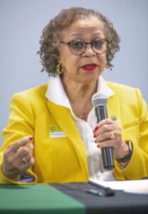 A woman with curly hair speaks into a microphone, wearing a bright yellow blazer and glasses.