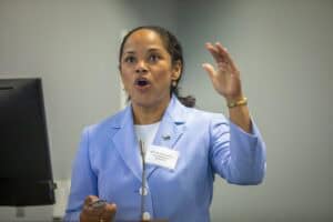 A woman in a light blue blazer passionately speaks while gesturing with her hand. There's a microphone on the stand.