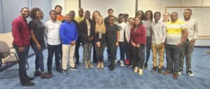 A diverse group of individuals poses together in a classroom setting, smiling and interacting.