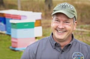A smiling man in a gray shirt and army cap stands in front of colorful beehives outdoors.