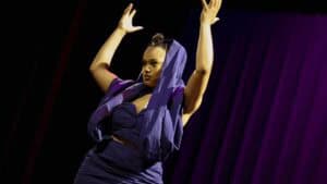 A woman performing a dance move on stage with purple curtains in the background.