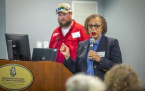 A woman speaking into a microphone at a conference with a man in a red shirt in the background.