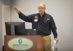 A man gestures while speaking at a podium with a computer, promoting agricultural research.