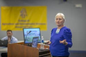 A woman in a blue top presents at a conference, gesturing towards a computer screen with information.