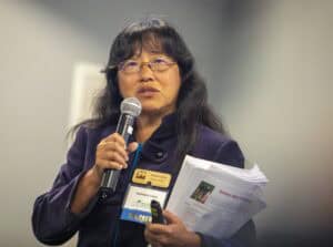 A woman with long dark hair and glasses speaks into a microphone while holding documents. She wears a dark jacket and has conference badges on her chest, standing in front of a blurred background.