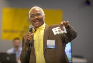 A speaker passionately addresses an audience while holding a microphone and materials in hand.