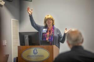 A woman gestures enthusiastically while speaking at a podium in a classroom setting.