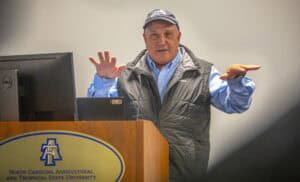 A speaker gestures while presenting at a university event, with a microphone and laptop in front of them.