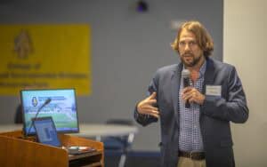 A man speaks into a microphone while gesturing, with a presentation screen nearby and a banner in the background.