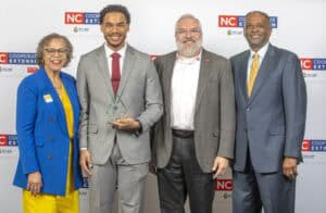 Four individuals stand together, smiling, with one holding an award. They are dressed in formal attire against a…
