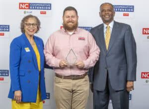 Three individuals stand together, one holding an award, in front of a backdrop featuring logos for NC Cooperative…