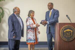 A man in a suit hands a plaque to a woman in a colorful coat, while another man looks on.