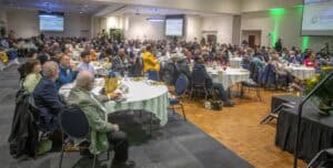 Attendees at a conference or seminar in a large, well-lit hall with presentation screens and round t.