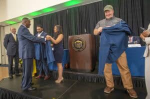 High-level award ceremony with individuals on stage, one person receiving a jacket, others presentin.