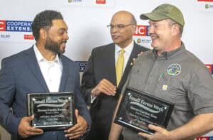 Two men stand holding awards, smiling at a third man in a suit who gestures between them.