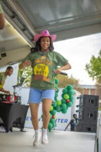 A woman dancing on stage wearing a pink cowboy hat and green "Hoggles Out West" t-shirt at an outdoo.