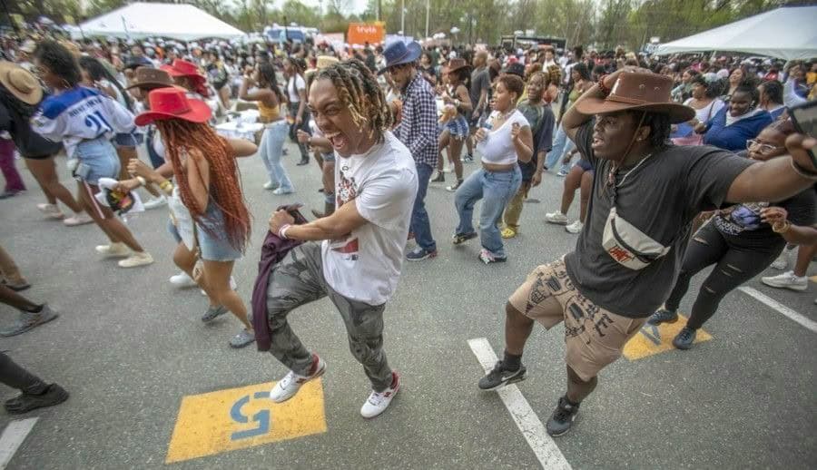 Crowd dancing joyfully at an outdoor event, with diverse participants in casual attire and hats.