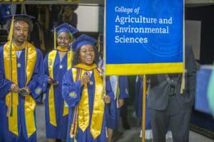 Graduates in blue caps and gowns celebrate, holding a banner for the College of Agriculture and Environmental Sciences.